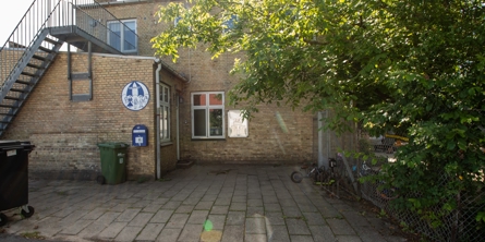 Indgangspartiet til daginstitutionen Børnely i Næstved Kommune. Bygningen er et gammelt KTAS  hus i gule sten. På højre side ligger legepladsen med grønne buske der vokser ud over hegnet.