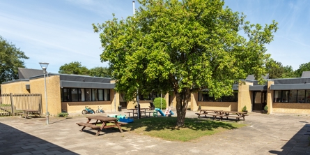 Indgangspartiet til daginstitutionen Eventyrgården i Næstved Kommune. Bygningen er opført i lyse sten og taget er fladt. Foran bygningen er et stort træ, med bænke nedenunder.