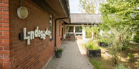 Indgangspartiet til daginstitutionen Lindegården i Næstved Kommune. Bygningen er opført i røde mursten. Der er en flisegang  langs huset. På højre side ligger  en lille parklignende have.