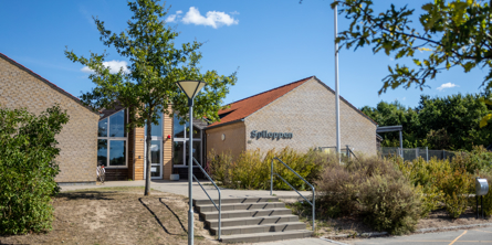 Indgangspartiet til daginstitutionen Spiloppen i Næstved Kommune. Der er en mindre trappe op til indgangspartiet. Indgangen har et stort glasparti. Der vokser træer og buske rundt om huset.