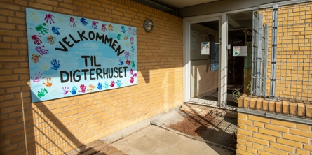 Indgangspartiet til daginstitutionen Digterhuset  i Næstved Kommune. Huset er opført i gule mursten. På muren hænger et stort blåt velkomstskilt. 