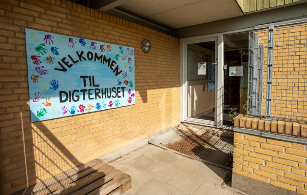 Indgangspartiet til daginstitutionen Digterhuset i Næstved Kommune. Huset er opført i gule mursten. På muren hænger et stort blåt velkomstskilt.