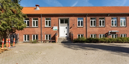 Indgangspartiet til daginstitutionen Frit 16 i Næstved Kommune. Huset et en stort rød stens bygning i to plan. Der vokser lave buske til højre foran huset og stor træer til venstre.