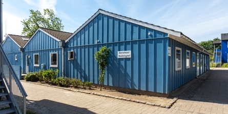 Indgangspartiet til daginstitutionen Nøddehegnet i Næstved Kommune. Bygningen er i blåt træ og der er belagt med fliser rundt om hele bygningen.   