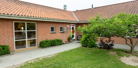 Indgangspartiet til daginstitutionen Evigglad i Næstved Kommune. Bygningen er et vinkelhus opført i røde mursten. Foran huset er der græsplæne med mindre træ og buske. 