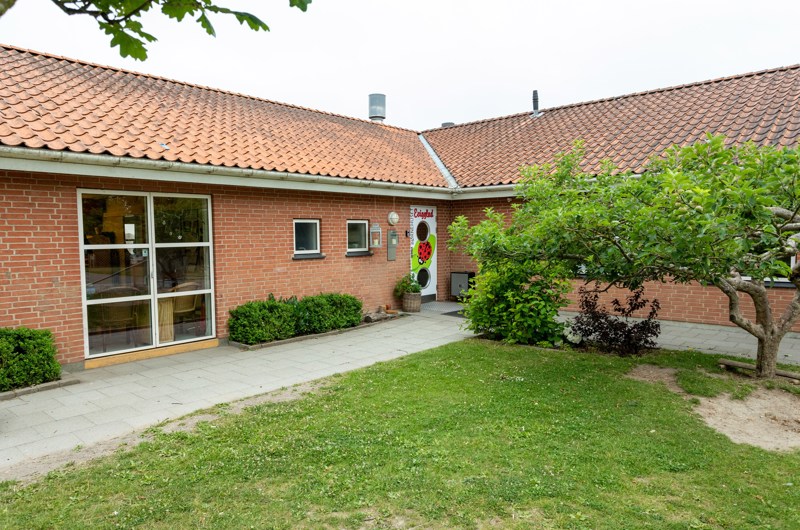 Indgangspartiet til daginstitutionen Evigglad i Næstved Kommune. Bygningen er et vinkelhus opført i røde mursten. Foran huset er der græsplæne med mindre træ og buske.