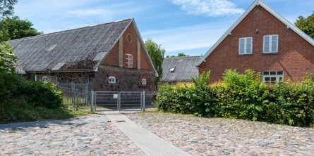 Indgangspartiet til daginstitutionen Sognefogedgården i Næstved Kommune. Bygningen er den gamle Sognefogedgård som er opført i røde mursten. langs indgangen ligger brosten og fliser. Bygningen er omkredset af grønne buske og hæk. 