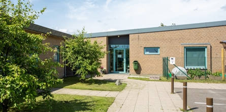 Indgangspartiet til daginstitutionen Tappernøje Børnehus i Næstved Kommune. Huset er en nyere bygning opført i røde mursten. til højre for indgangspartiet ligger parkeringspladsen. På venstre side vokser der små træer, på små græsarealer.  