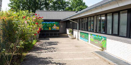 Indgangspartiet til daginstitutionen Mælkebøtten i Næstved Kommune. Huset er bygget i hvide mursten. På ydrevæggene  er der opsat plader med børnetegninger på. Der er rosenbuske langs indgangspartiet.