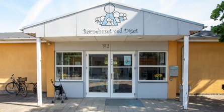 Indgangspartiet til daginstitutionen Diget i Næstved Kommune. Husets facade er gul. Indgangen er overdækket og der er vinduer og døre i glas. 