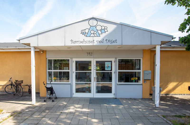 Indgangspartiet til daginstitutionen Diget i Næstved Kommune. Husets facade er gul. Indgangen er overdækket og der er vinduer og døre i glas.