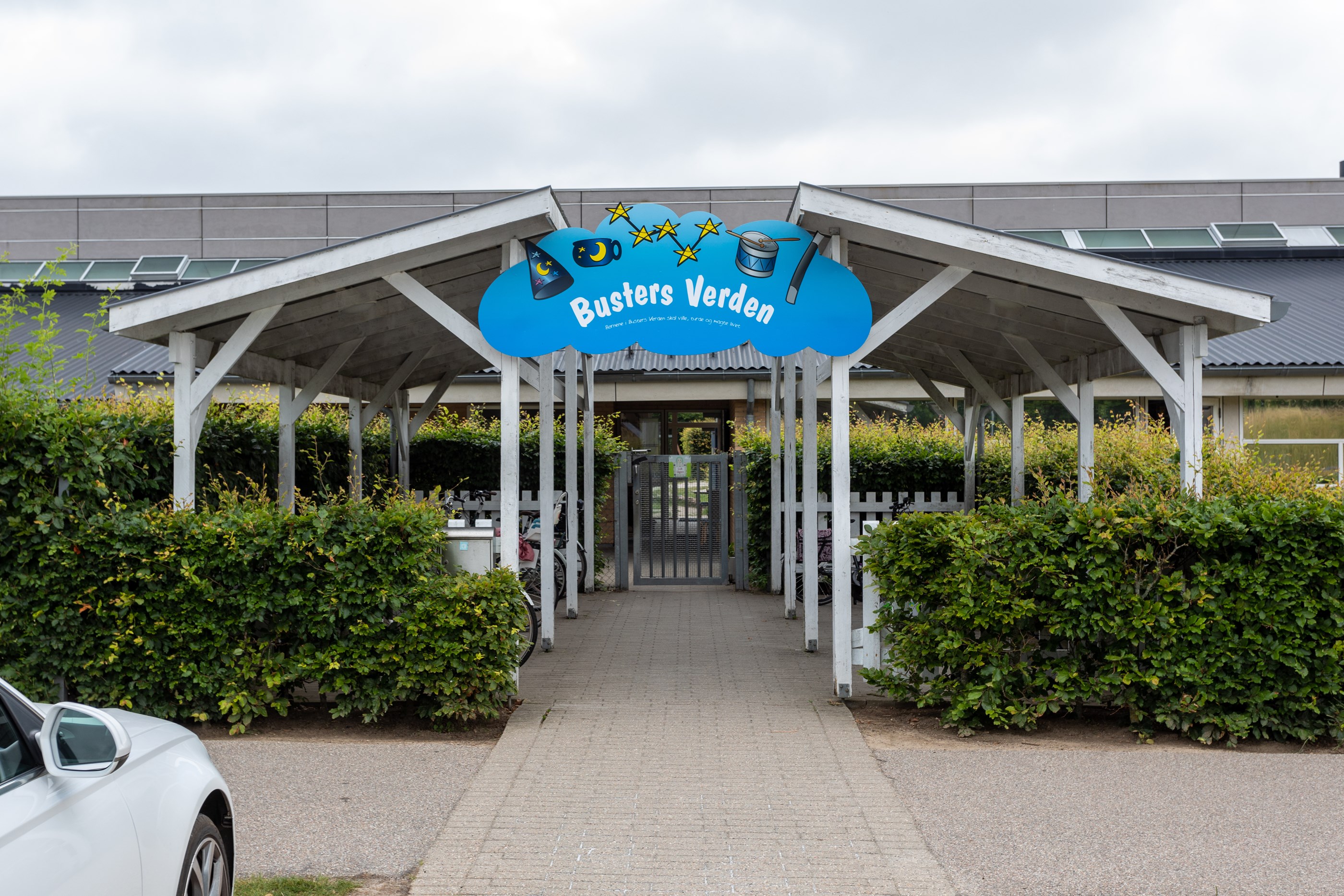 Indgangspartiet til daginstitutionen Busters Verden i Næstved Kommune. Over indgangen er der et stort blåt skilt med institutionens navn, samt grønne hække langs indgangen.