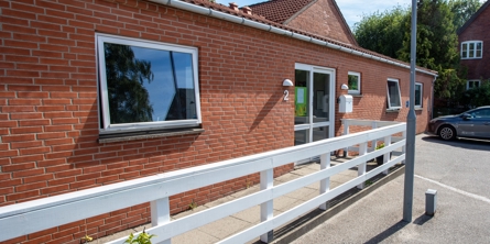 Indgangspartiet til daginstitutionen Lillevang i Næstved Kommune. Huset er en lang et plans bygning i røde mursten. Foran er et hvidt lavt træhegn, der løber langs bygningen. 