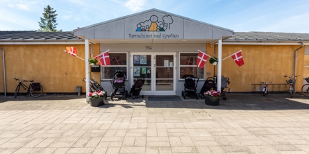 Indgangspartiet til daginstitutionen Grøften i Næstved Kommune. Husets facade er gul. Indgangen er overdækket og der er vinduer og døre i glas. Der er sat dannebrogsflag op på stolperne til indgangspartiet.  
