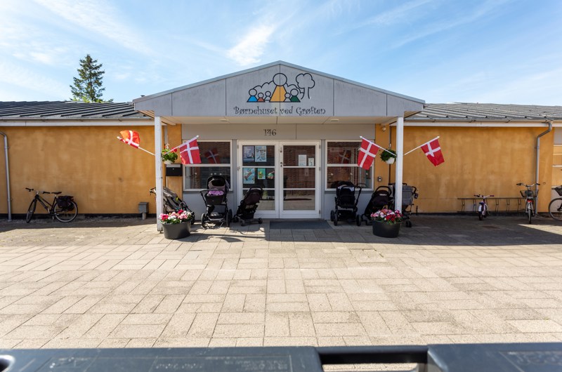 Indgangspartiet til daginstitutionen Grøften i Næstved Kommune. Husets facade er gul. Indgangen er overdækket og der er vinduer og døre i glas. Der er sat dannebrogsflag op på stolperne til indgangspartiet.