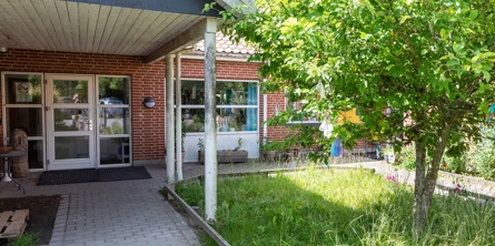 Indgangspartiet til daginstitutionen Eventyrhuset i Næstved Kommune. Huset er bygget i røde mursten. Indgangen er overdækket og der er en lille græsplæne, hvor der er plantet et træ. 