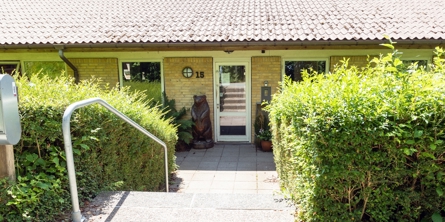 Indgangspartiet til daginstitutionen Tjørnehuset i Næstved Kommune. Bygningen er i et plan og bygget i gule mursten. Der er en mindre trappe ned til indgangsdøren. Der vokser hæk på begge sider af trappen. 