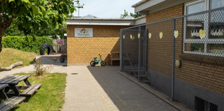 Indgangspartiet til daginstitutionen Manøhytten i Næstved Kommune. Bygningen er opført i gule mursten. Der løber et trådhegn langs bygningen. Overfor hegnet er der et græsareal med bord bænke sæt.  