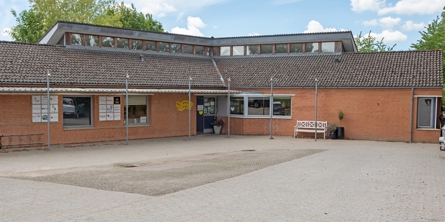 Indgangspartiet til daginstitutionen Ellebæk i Næstved Kommune. Huset er et stort vinkelhus med ovenlysvinduer. Bygningen er opført i røde mursten. Indgangen ligger ud til parkeringspladsen.  