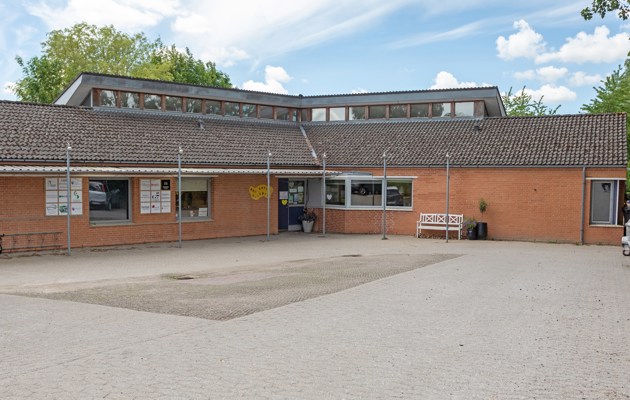 Indgangspartiet til daginstitutionen Ellebæk i Næstved Kommune. Huset er et stort vinkelhus med ovenlysvinduer. Bygningen er opført i røde mursten. Indgangen ligger ud til parkeringspladsen.